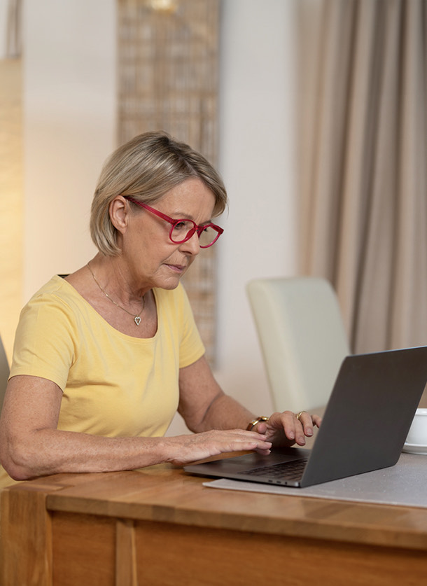  The image depicts an older woman sitting at her living room table, working on a grey laptop. She wears a yellow short-sleeved top and red reading glasses, with short blonde hair visible. The indoor setting features light curtains in the background, and part of another chair is visible. The red reading glasses add a pop of color to the scene, creating a cozy and focused atmosphere. 
