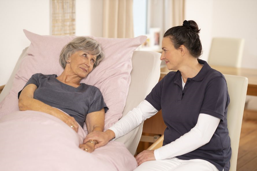 Eine Pflegerin hält die Hand von einer älteren Dame, die im Bett liegt