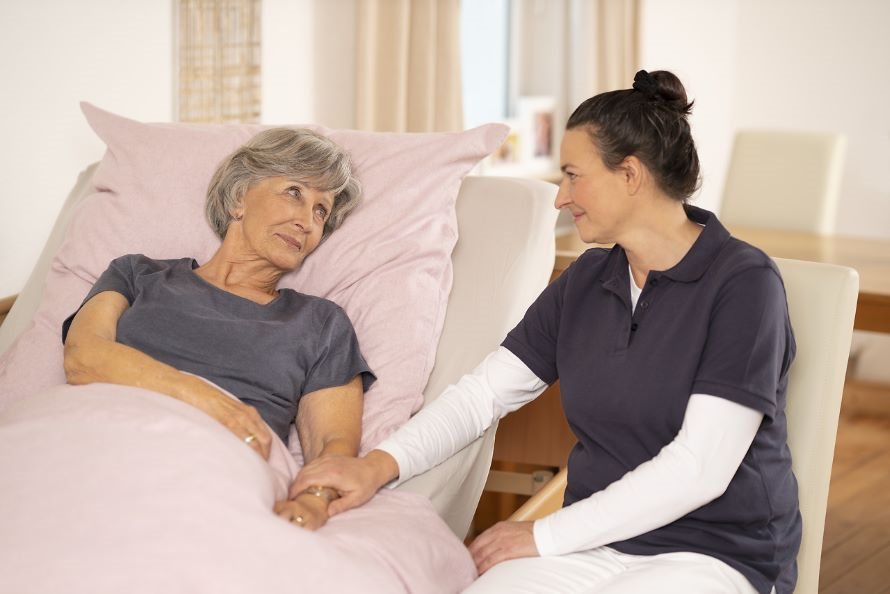 Eine Pflegerin hält die Hand von einer älteren Dame die im Bett liegt